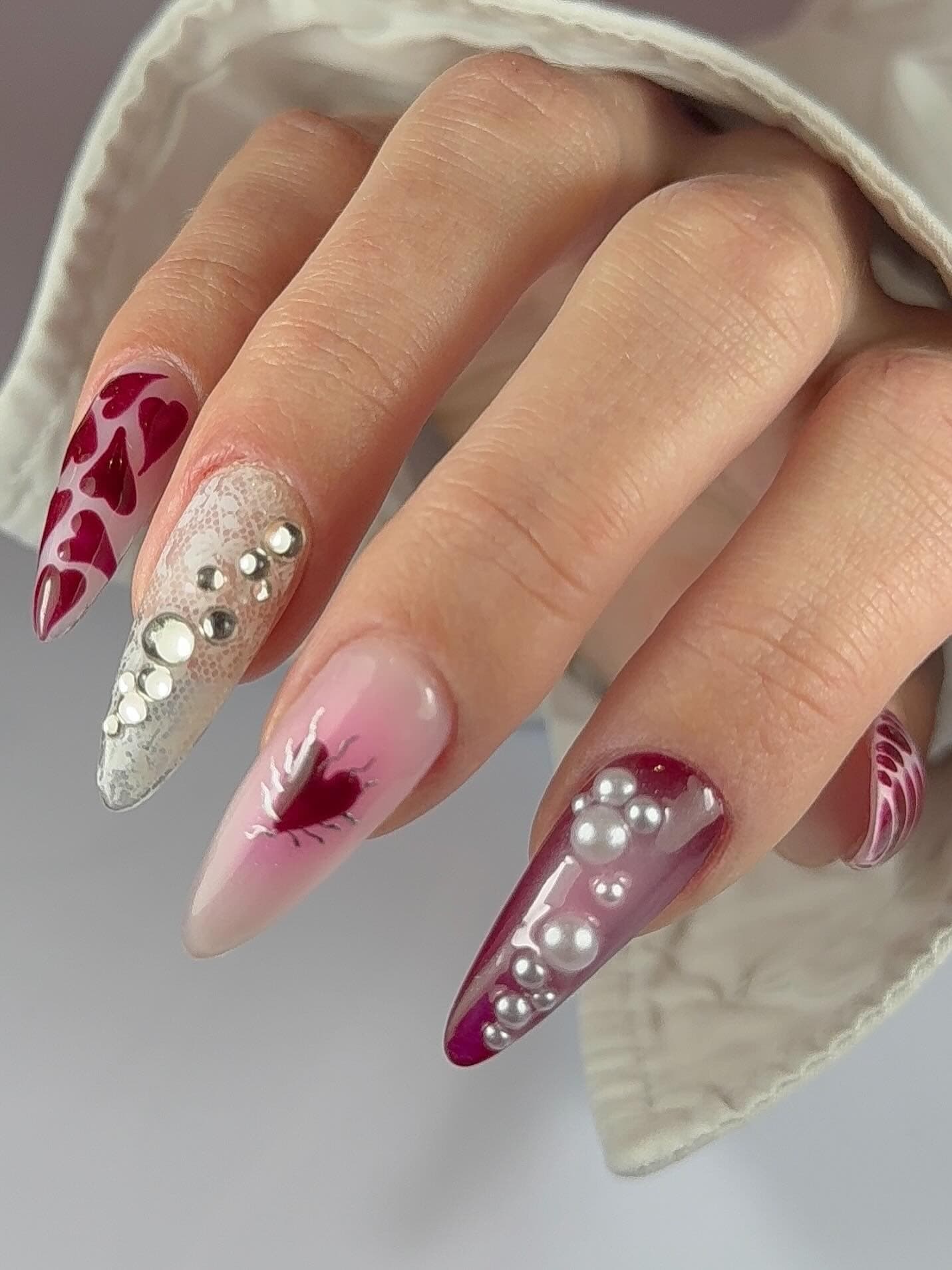 Nail art en amande mêlant cœurs bordeaux, strass argentés et perles.